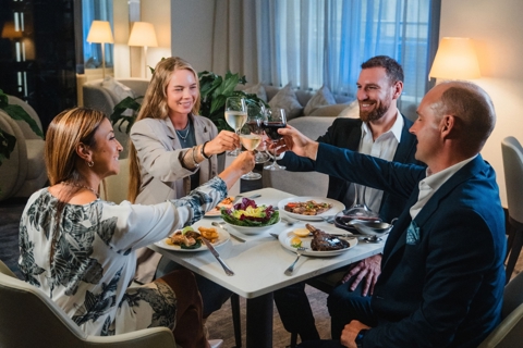 Four individuals are enjoying a meal together at a restaurant. They are seated around a square table with dishes of food and are in the midst of a toast, with their wine glasses raised. The atmosphere appears convivial and celebratory.